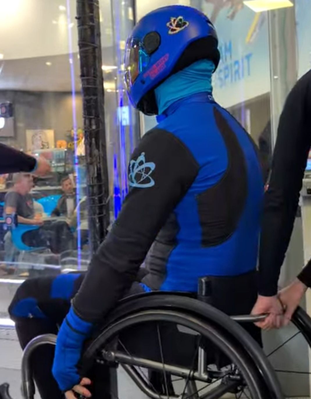 A flyer in a blue flight suit and helmet sits in a wheelchair at the entrance to an indoor wind tunnel while a helper steadies the chair.