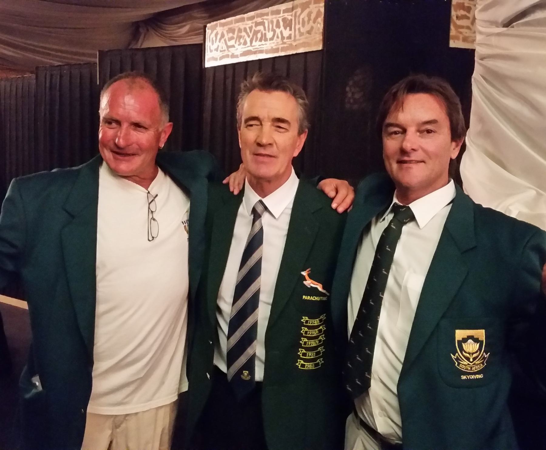 Three men with their arms around each other, wearing green South African team blazers. The centre blazer carries a springbok badge, the word Parachuting and the years 1974/5, 1976/7, 1978/9, 1981 and 1983. The blazer on the right carries a South Africa Skydiving crest.